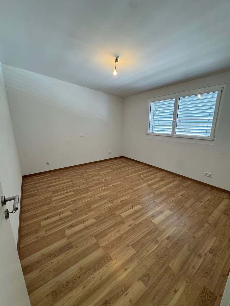 The image shows a spacious bedroom with a hardwood floor, white walls, and a single window with blinds. The room appears to be well-lit with a ceiling light fixture.