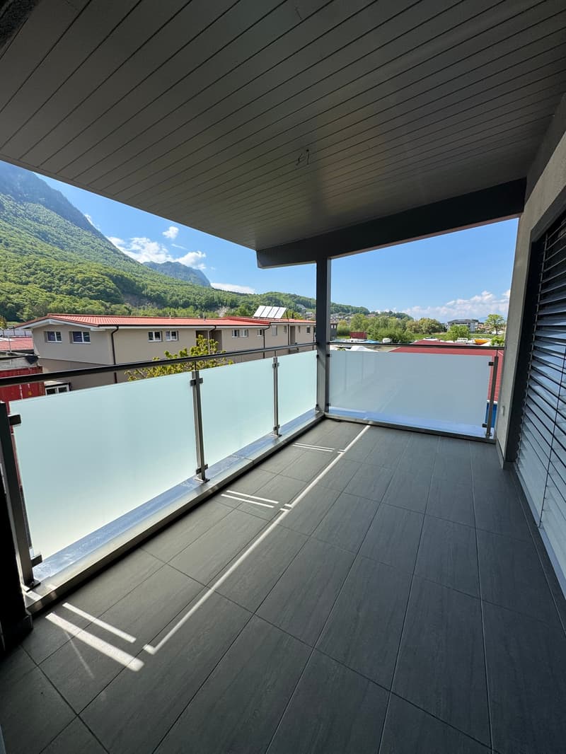 Covered balcony with glass railing, overlooking a residential area with mountains in the background. The balcony has a tiled floor and a wooden ceiling.