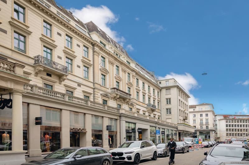 This image shows a multi-story building with a classical architectural style, featuring ornate balconies, columns, and decorative elements. The building has several retail stores on the ground floor, including a Gucci store. There are cars parked on the s