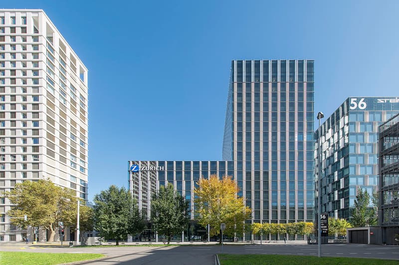 Modern office building with glass facade, multiple floors, and a sign reading 'ZURICH'. Adjacent to a high-rise residential building. Trees and greenery in the foreground. Clear blue sky.