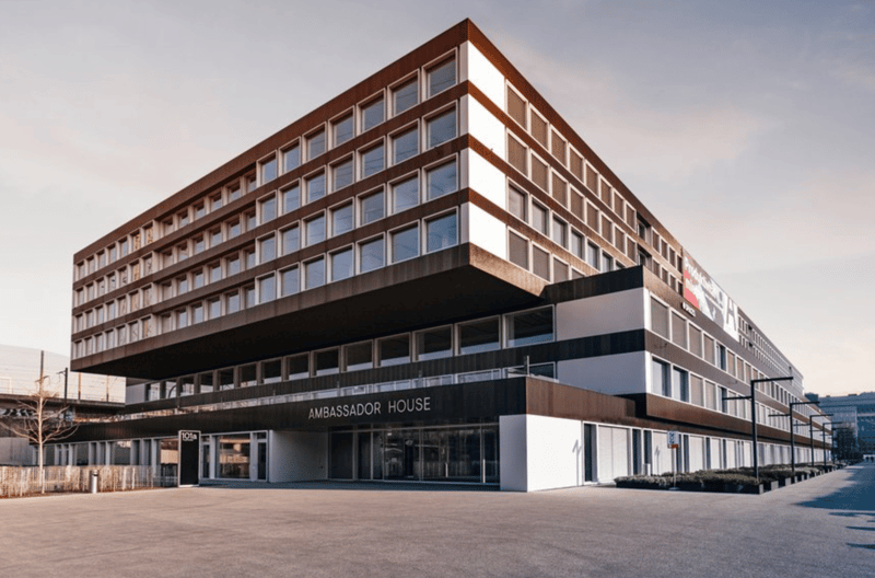 Modern office building, multiple floors, glass windows, entrance with glass doors, name board 'AMBASSADOR HOUSE', parking area in front
