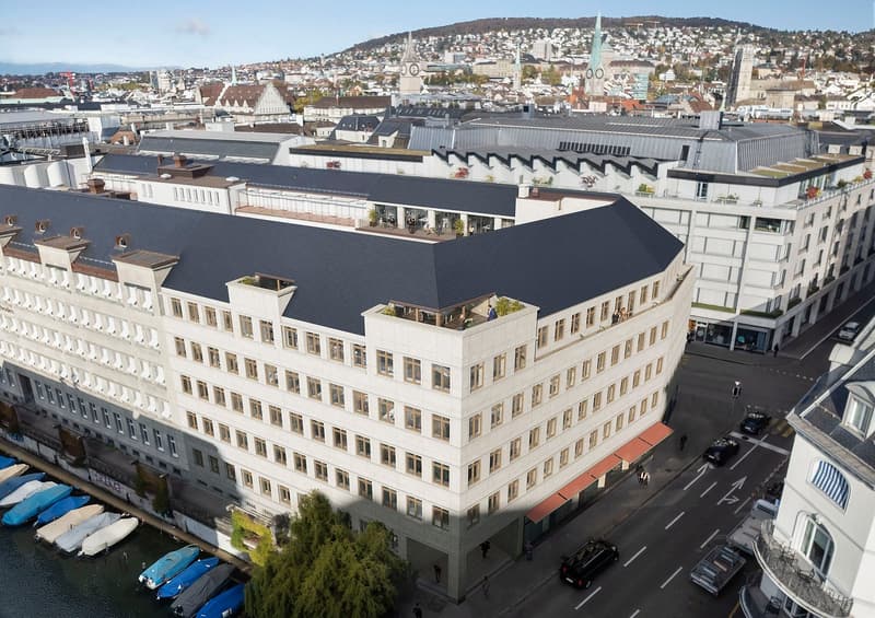 Modern office building with multiple floors, white exterior, black roof, multiple windows, balcony on the top floor, located in an urban area with a river and boats nearby.