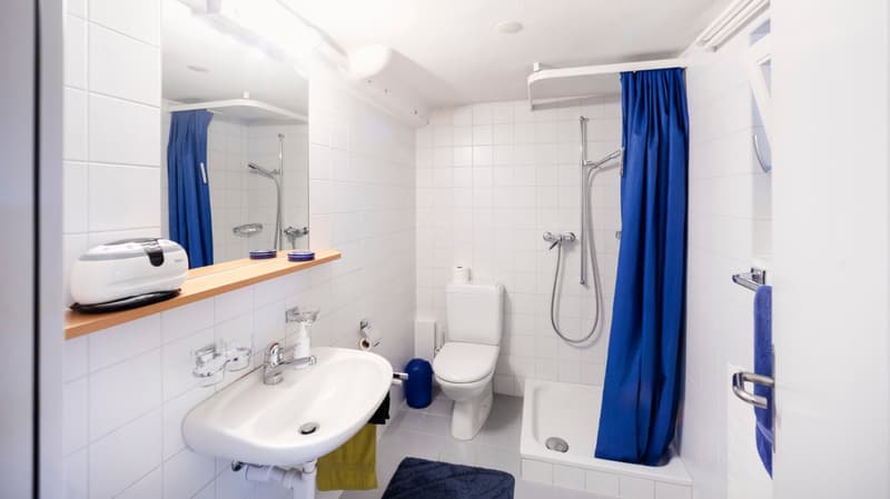 white tiles, white sink, white toilet, shower with blue curtain, mirror, blue towel