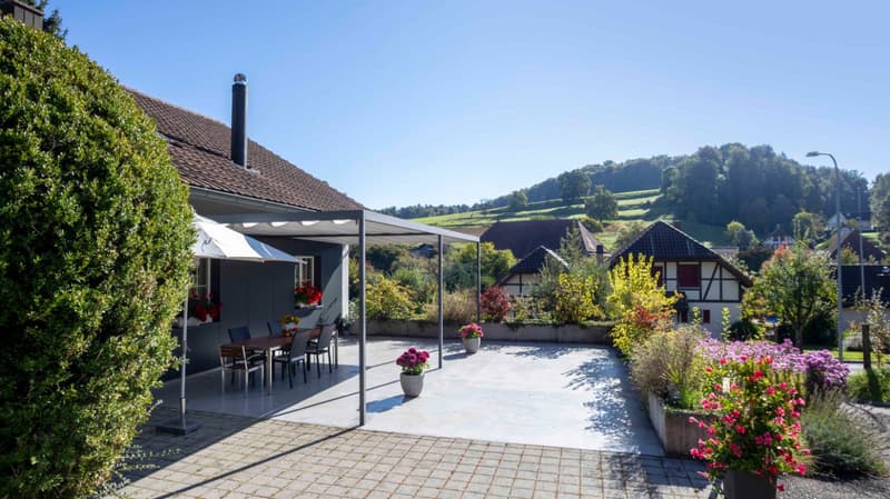Outdoor dining area with table and chairs, covered by a pergola, surrounded by plants and flowers, paved patio, and a view of the mountains.