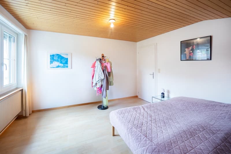 Bedroom with wooden floor, white walls, bed, clothes rack, picture frames, window with curtains, radiator, bedside table with lamp, and ceiling light.