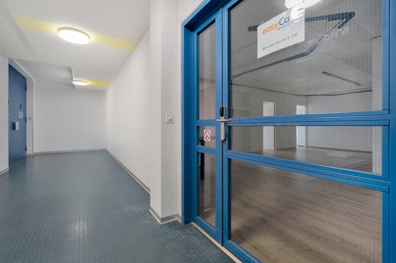 The image shows a hallway in a medical facility. The hallway has white walls and a tile floor. There is a blue-framed glass door with the 'Easy Co. Medical' logo visible through it. The hallway appears to be clean and well-lit.