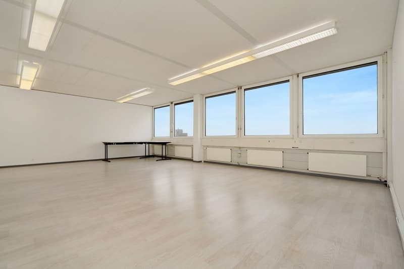 Large open room with white walls, hardwood floors, and large windows providing natural light. The room has several tables and chairs, suggesting it is used as an office or meeting space.