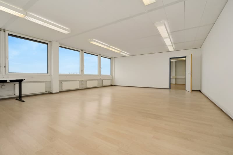 Large open office space with white walls, hardwood floors, and multiple windows providing natural light. The room has a piano and appears to be set up for office or commercial use.
