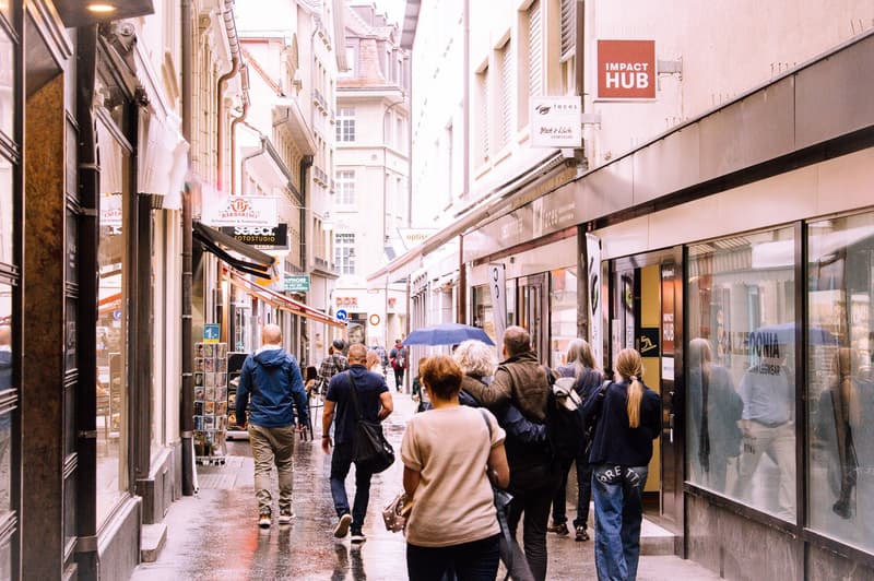 a narrow street with shops, people, buildings with glass windows and awnings, wet street from rain