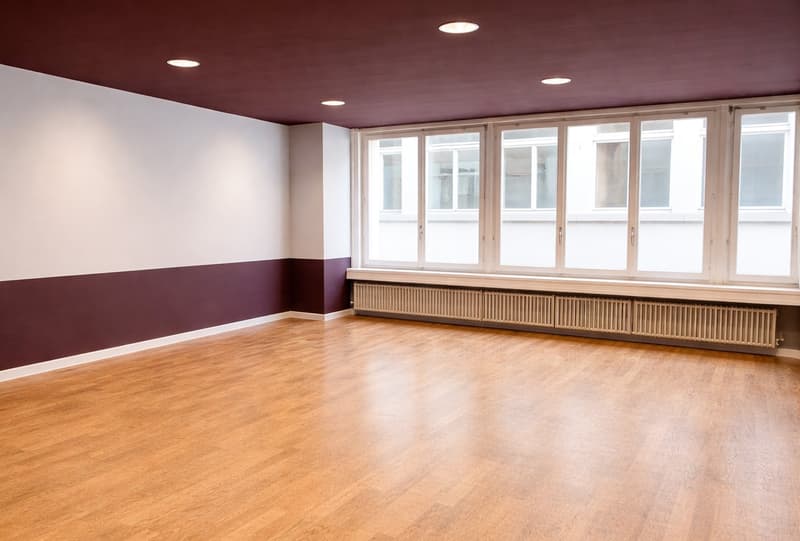 wide empty room, wooden floors, white and burgundy walls, large windows, and radiators