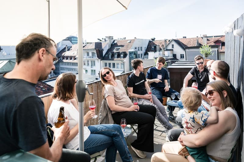 Group of people socializing on a balcony, wine glasses, baby, wooden barrier, plants, distant buildings.