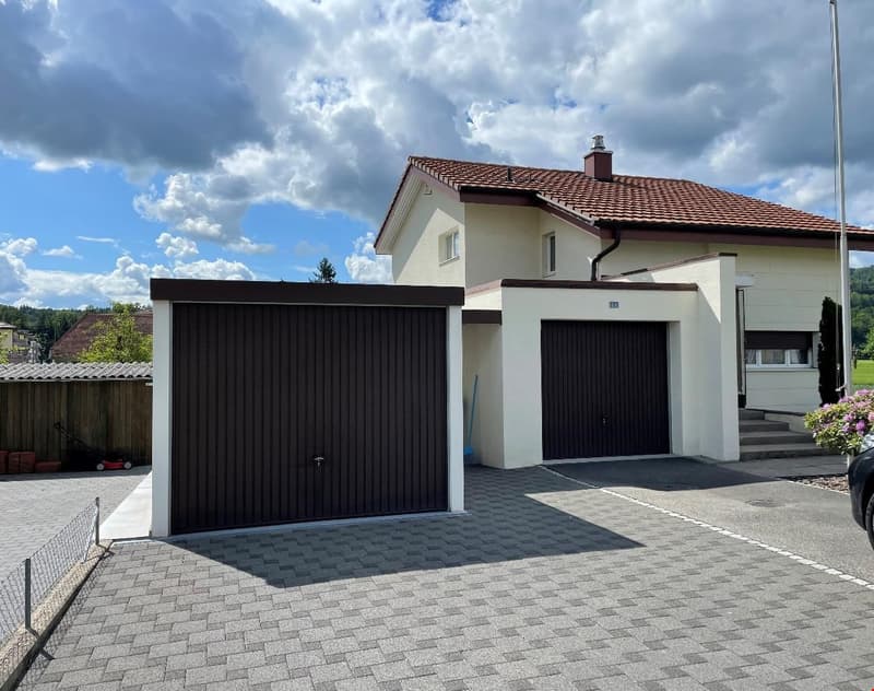 2-story house with white exterior walls, red tile roof, and black garage doors. The house has a paved driveway and a small garden area in the front.