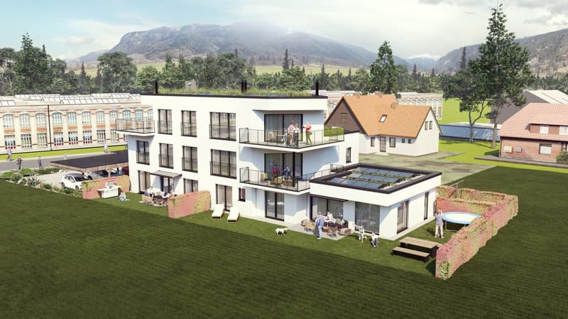 Multi-story apartment building with balconies, surrounded by green lawns and trees, with a parking area in the foreground. The building has a modern, glass-fronted design with a mix of white and wooden elements. In the background, there are mountains and