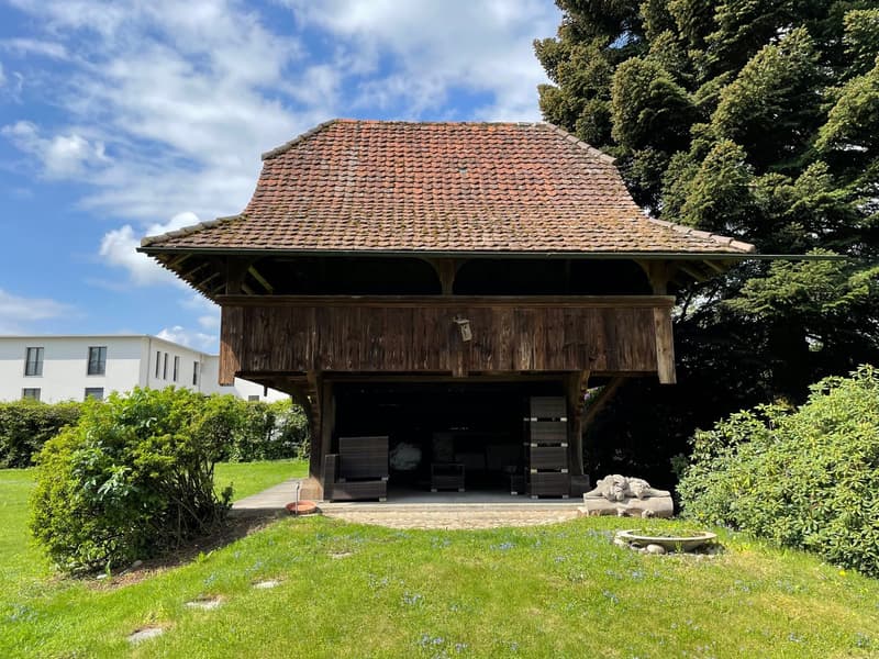 Old wooden house with red roof, wooden construction, surrounded by lush green grass and plants, with a white building behind it.