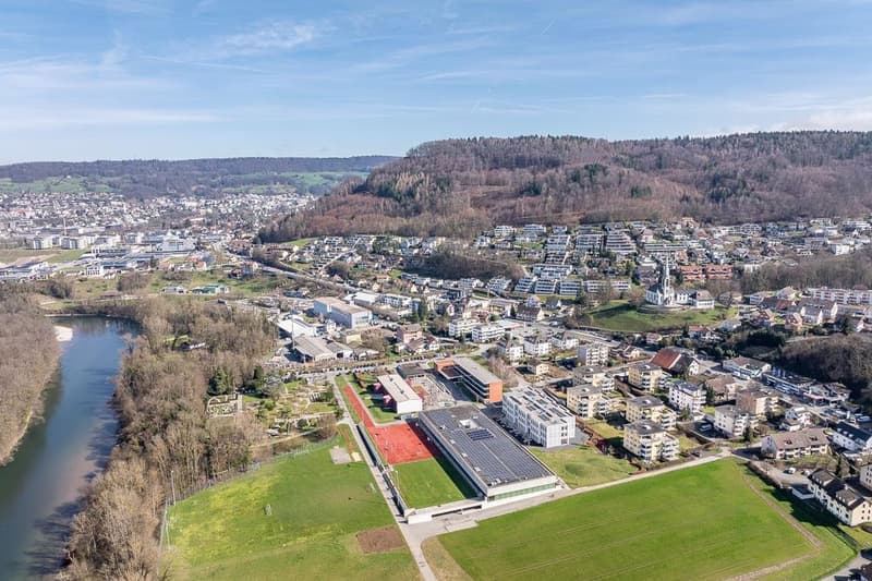 This image shows an aerial view of a town or city surrounded by hills and forests. The town has a mix of residential and commercial buildings, with some larger industrial or institutional buildings visible. There is a river or body of water running throug