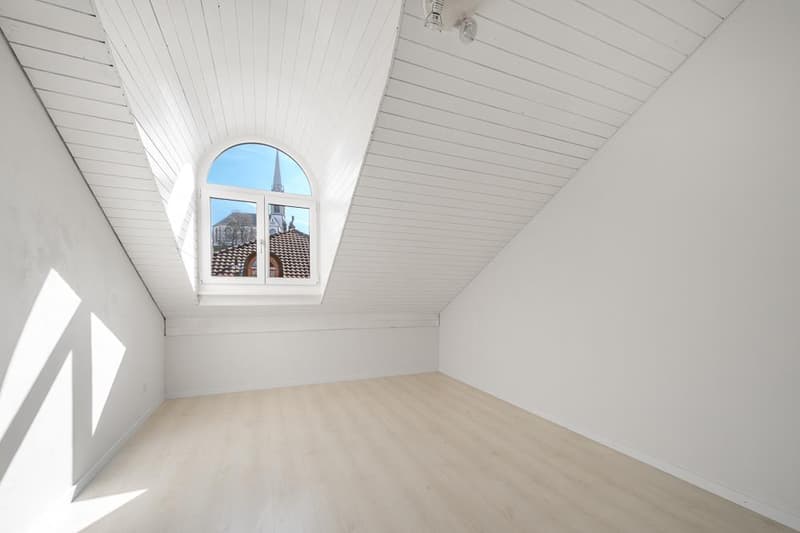 Spacious attic bedroom with white walls and ceiling, wooden floor, and a large arched window providing a view of the city skyline