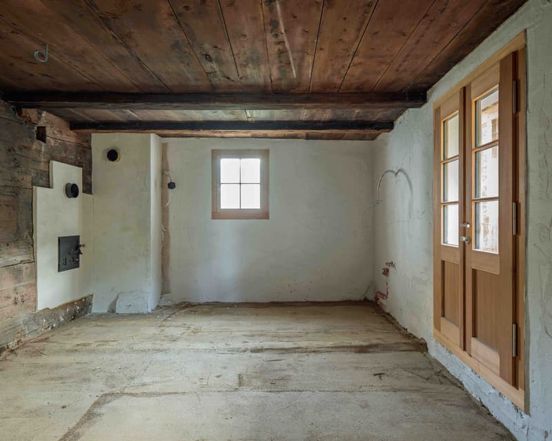 A room with wooden beams, white walls, concrete floor, a window, and a black fireplace on the wall.