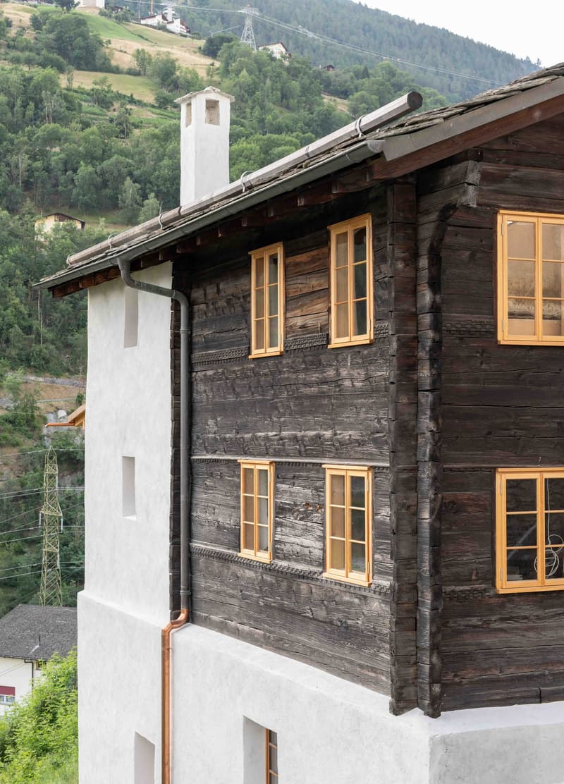 Detached house, rustic wooden facade, yellow window frames, white walls, sloped roof, chimney, mountains in the background.
