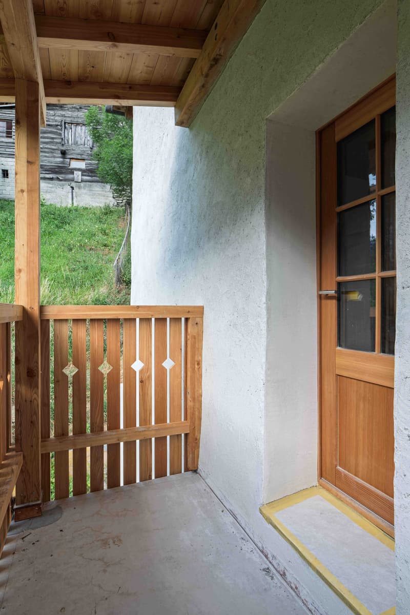 Wooden balcony, white walls, wooden railings, wooden door, concrete floor, yellow lines at the door entrance