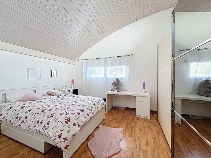 Bedroom with a bed, desk, and a rug. The bed has a floral bedspread. The desk has a stuffed toy and a lamp. The room has a wooden floor and white walls. There are two windows with white curtains.