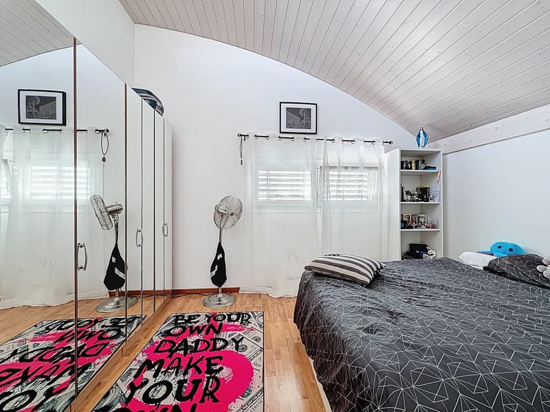 Bedroom with wooden floor, white walls, bed, rug, shelves, fans, curtains, and framed pictures.