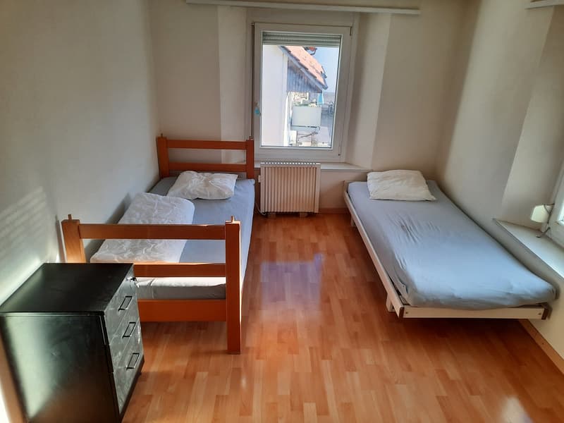 The image shows a bedroom with two single beds, a wooden dresser, and a window overlooking the exterior. The room has hardwood flooring and white walls.