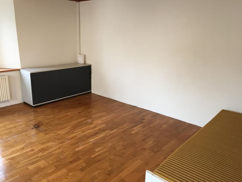 The image shows an empty room with a wooden floor and a white wall. There is a black cabinet or storage unit against the wall. The room appears to be a living room or bedroom based on the simple furnishings.