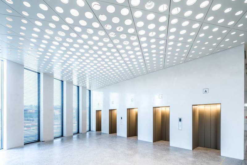 wide interior hall, white walls, gray floors, open elevator doors, windows with blinds