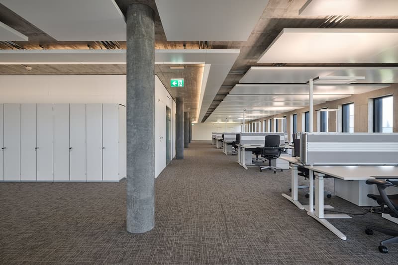 wide open space, grey floor, white walls, modern office chairs, desks, grey ceiling panels, glass windows on either side