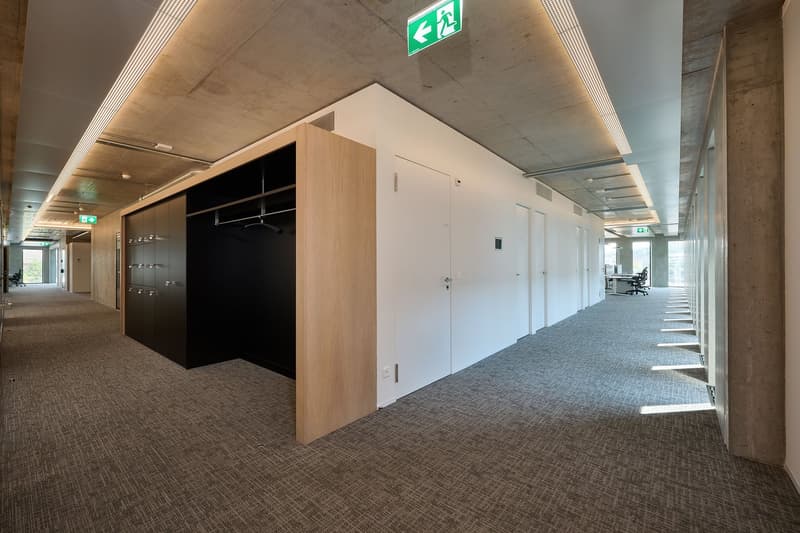 Long corridor with lockers, white walls, gray floor, doors, and emergency exit signs