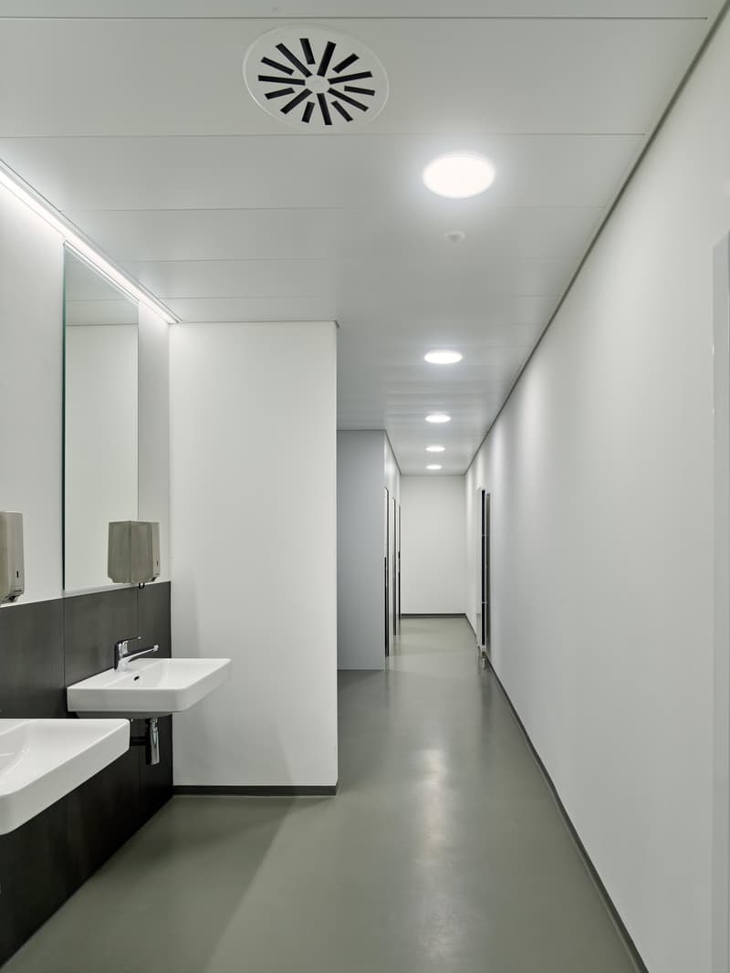 Empty corridor with white walls, gray floor, two sinks, two mirrors, and ceiling lights