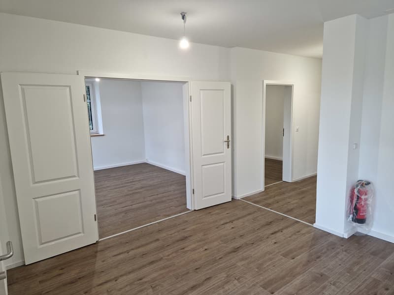 Empty room with white walls, wooden floor, three doors, and a fire extinguisher