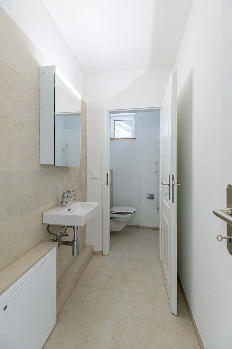 Modern bathroom with white tiles, a white vanity with sink, a mirror, and a toilet area with a small window.