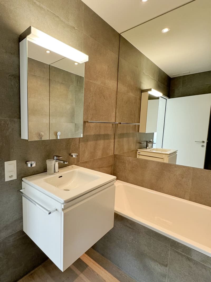 Modern bathroom with large mirror, bathtub, white sink, faucet, wooden floor, large light above the mirror