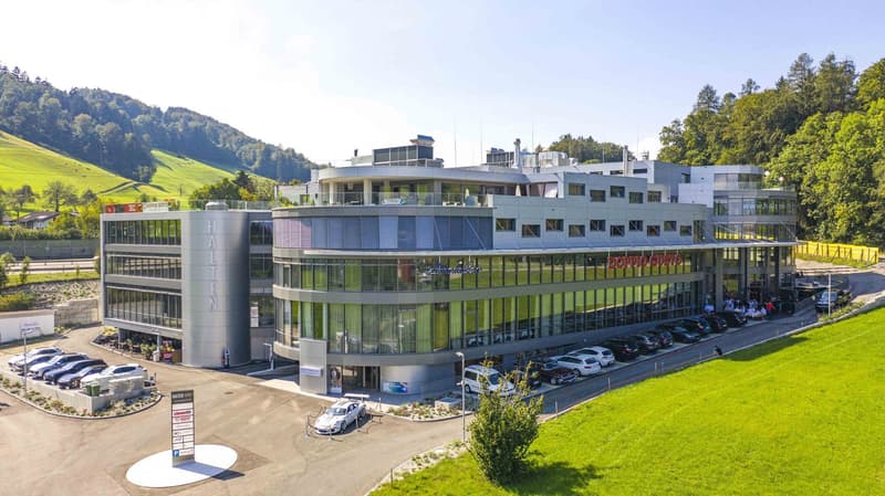 Modern building with glass facade, multiple floors, large parking area, surrounded by greenery