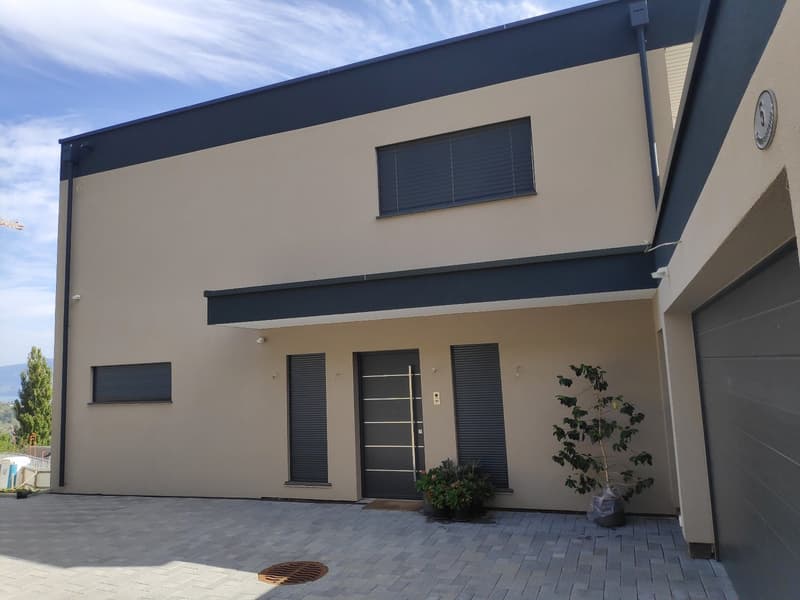 2-story modern house with beige exterior walls, black trim, and a garage door. The house has large windows and a paved driveway or entrance area.
