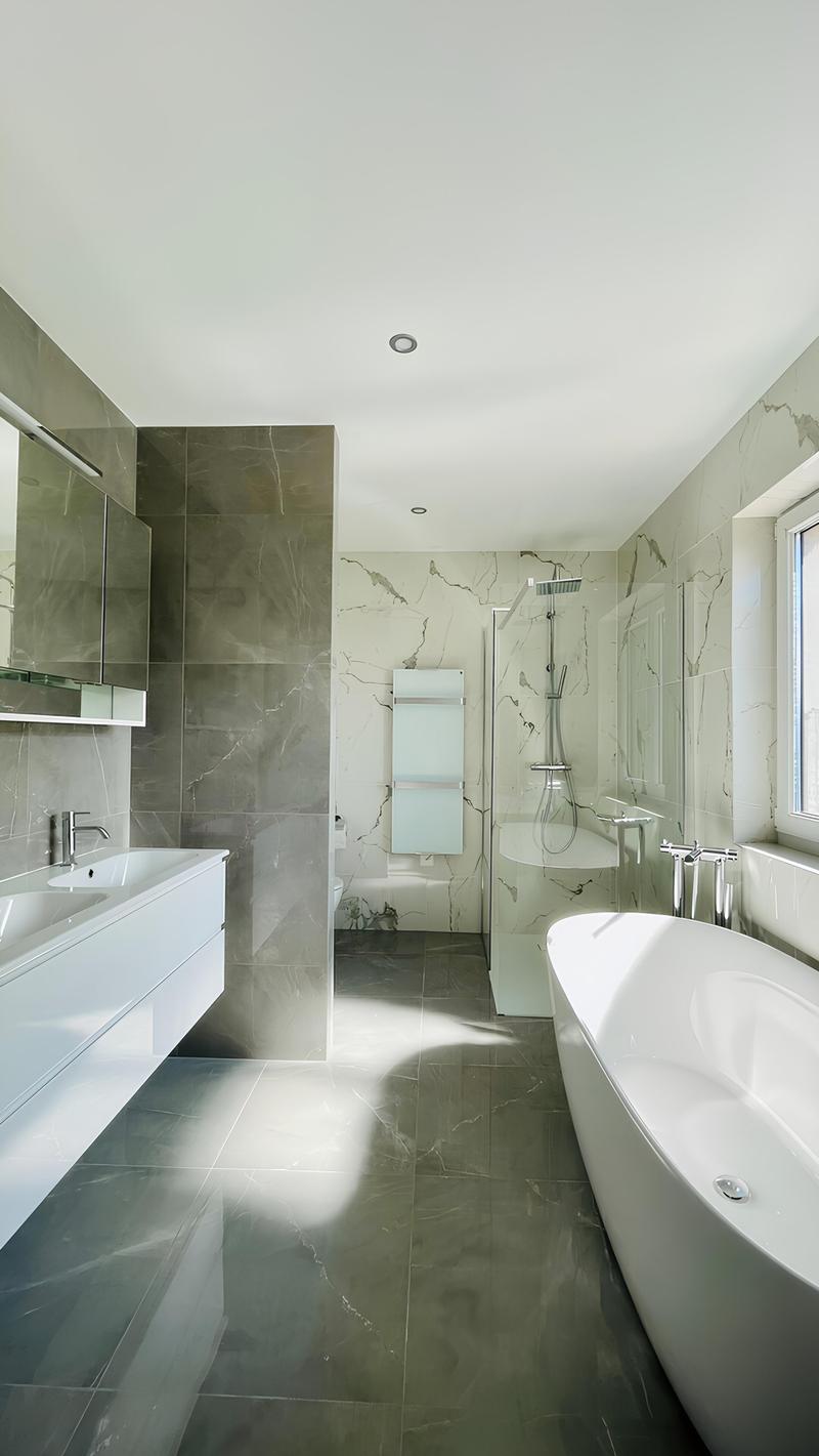 Spacious bathroom with grey marble-like tiles covering the walls and floor. The bathroom features a freestanding bathtub, a glass-enclosed shower area, and a vanity unit with a sink. The room has a modern and minimalist design.