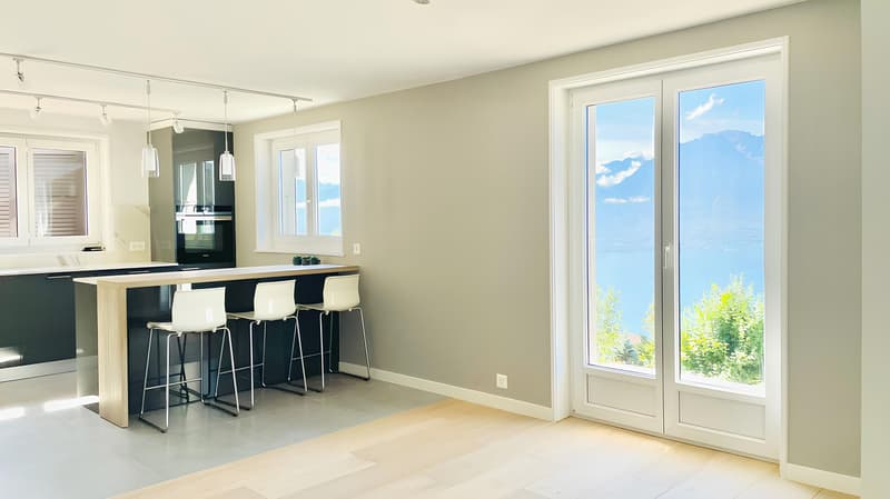 Spacious open-plan living room and kitchen with large windows offering a scenic view of the outdoors. The kitchen features a modern black and wood design with a breakfast bar and stools. The living area has a bright and airy feel with neutral-colored wall