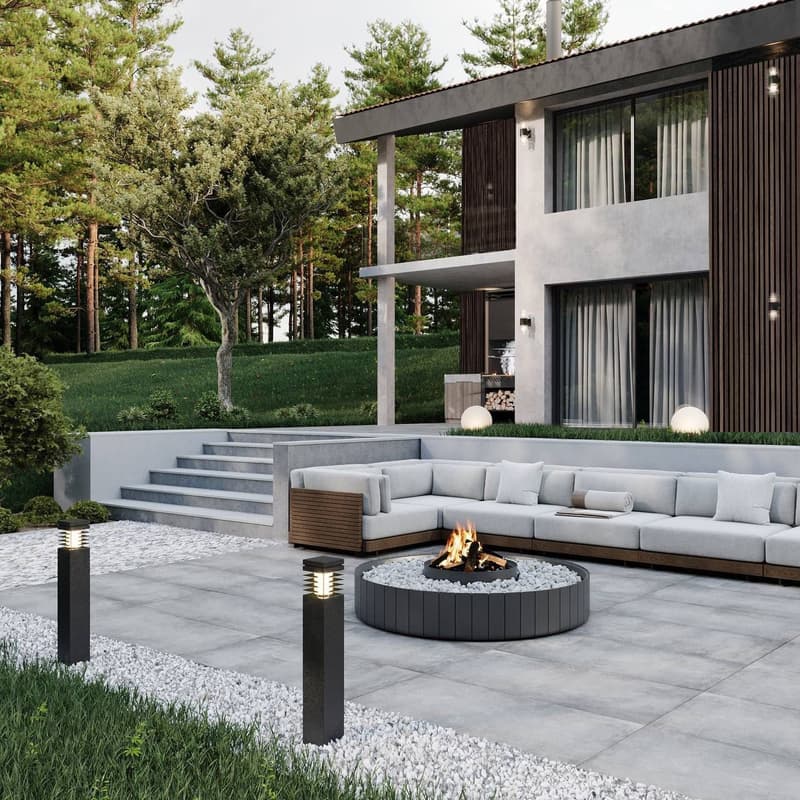 Modern outdoor living space with a large sectional sofa, a circular fire pit, and a raised patio with steps leading down to a grassy area surrounded by pine trees. The house has a sleek, contemporary design with large windows and a covered patio area.