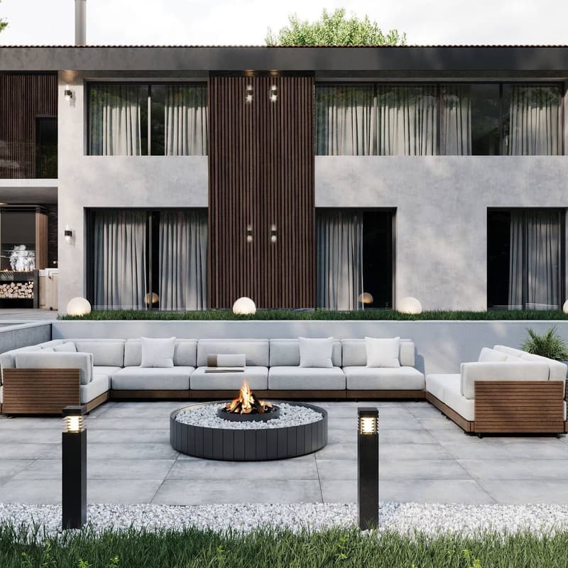 The image shows a modern and stylish outdoor living area. The space features a large sectional sofa with white cushions, a circular fire pit in the center, and a wooden deck with a grassy area surrounding it. The building in the background has a sleek, co