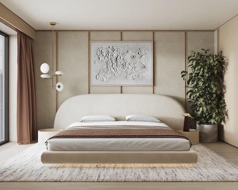 Spacious bedroom with a large, low-profile bed frame, neutral-toned walls and furnishings, a textured artwork above the bed, and a potted plant in the corner. The room also features a large window and curtains, as well as some lighting fixtures.