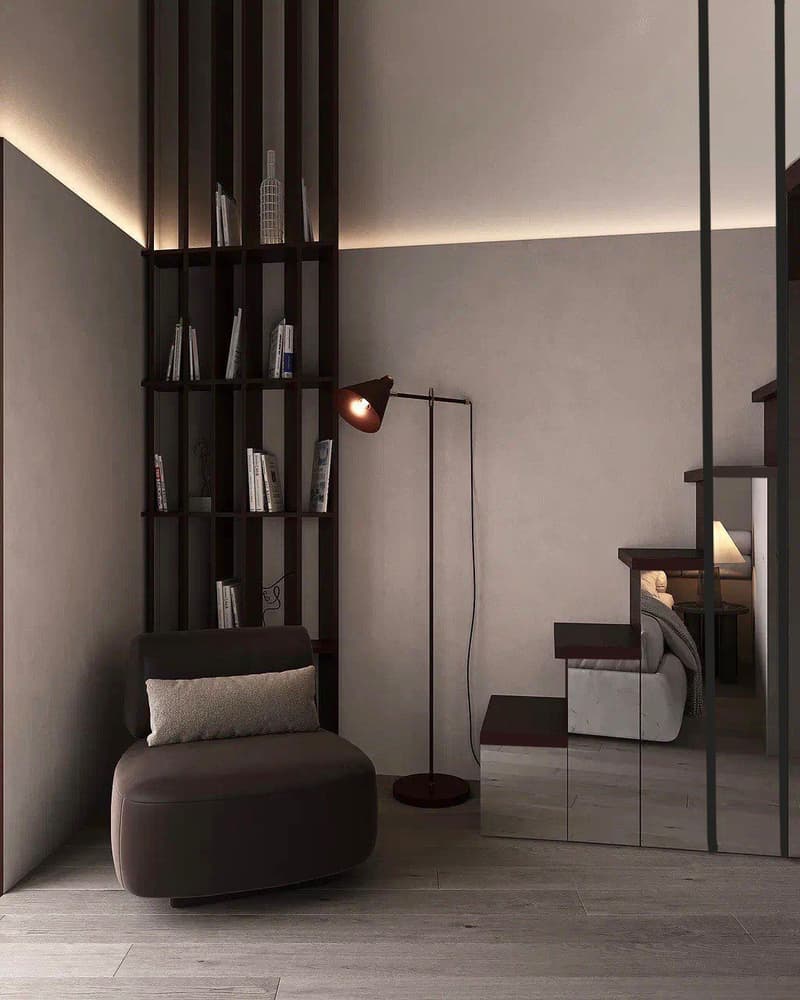The image shows a modern and minimalist living room with a dark color scheme. The room features a black bookshelf, a gray armchair, and a floor lamp. The floor is made of dark hardwood, and the walls are painted in a neutral gray tone. The room also has a
