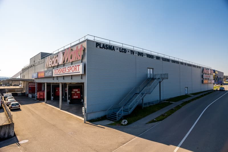 Large building with parking in front, stairs leading up to entrance, storefronts, and signage.