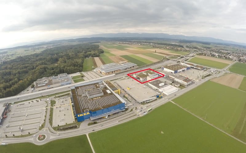 Industrial complex with a prominent IKEA store, surrounded by extensive green fields, trees, and a road network. The complex includes several buildings, with the IKEA store featuring a blue facade.