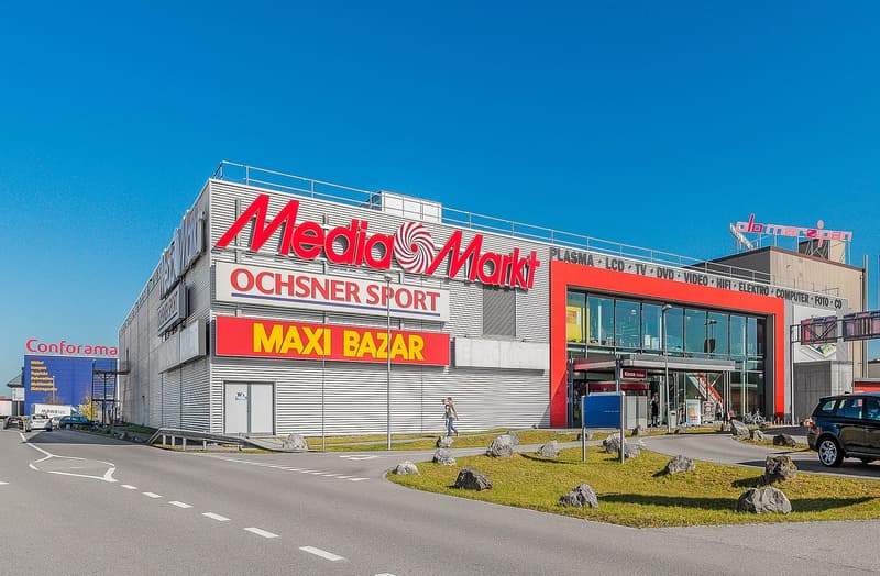 Retail complex with Media Markt, Ochsner Sport, and Maxi Bazar signage.