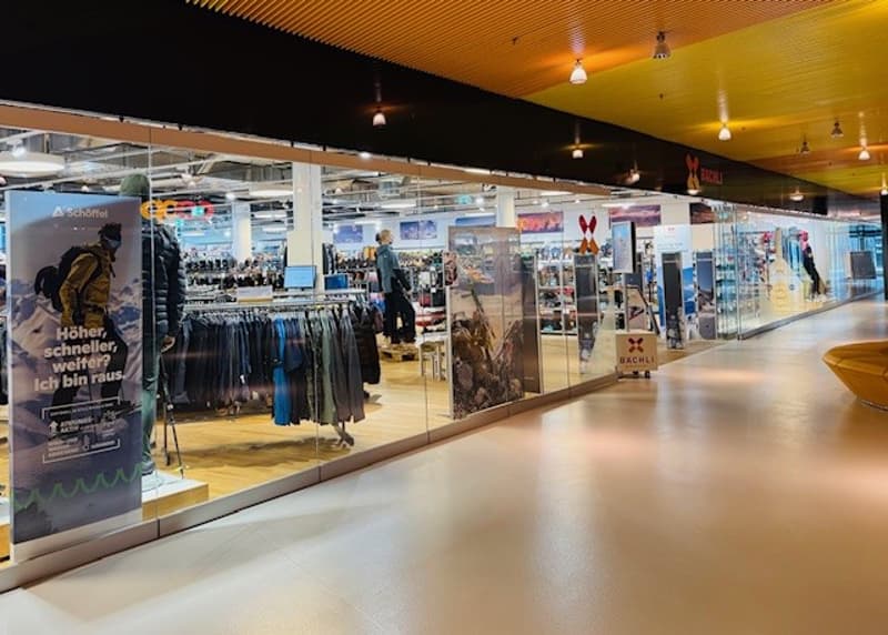 A clothing store with glass walls and doors, displaying various clothing items on hangers and shelves. The store has a modern design with a yellow ceiling and white walls. There are people inside the store, and some are walking outside. The store has a si