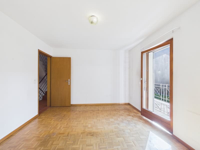 Empty room with wooden flooring, white walls, wooden door, glass door, and staircase leading to balcony with metal railing.