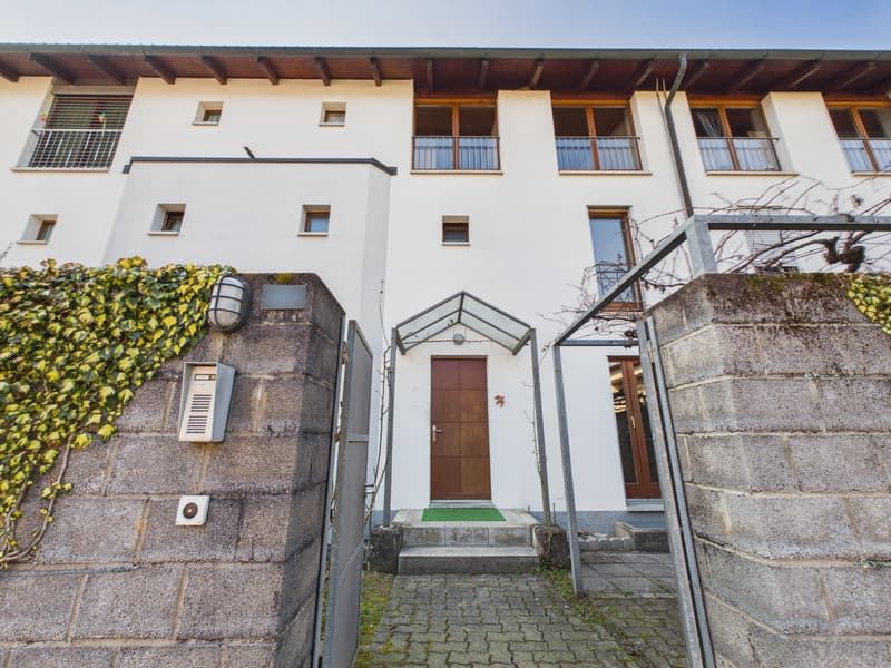 Exterior entrance with a wooden door, a pathway leading to it, and stone walls with attached ivy and a lamp. The house has a white exterior with multiple windows and balconies.