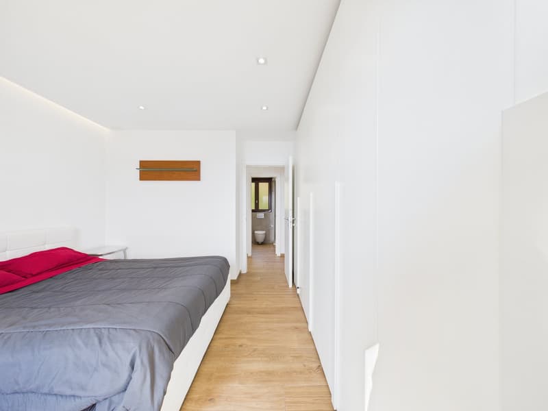 Bedroom with wooden floor, white walls, bed with gray sheets, red pillows, small table, hallway leading to bathroom