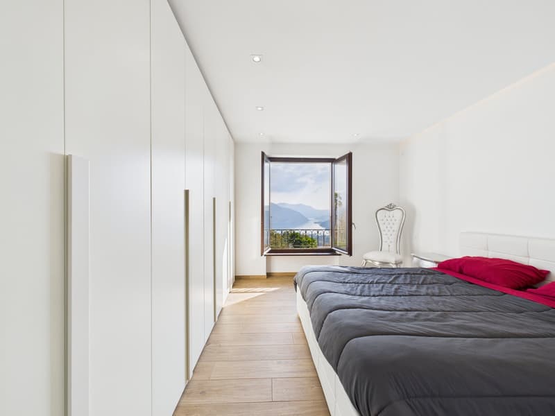 modern bedroom with white walls, wooden floor, large window with balcony, bed with gray sheets and red pillows, white chair, built-in wardrobes